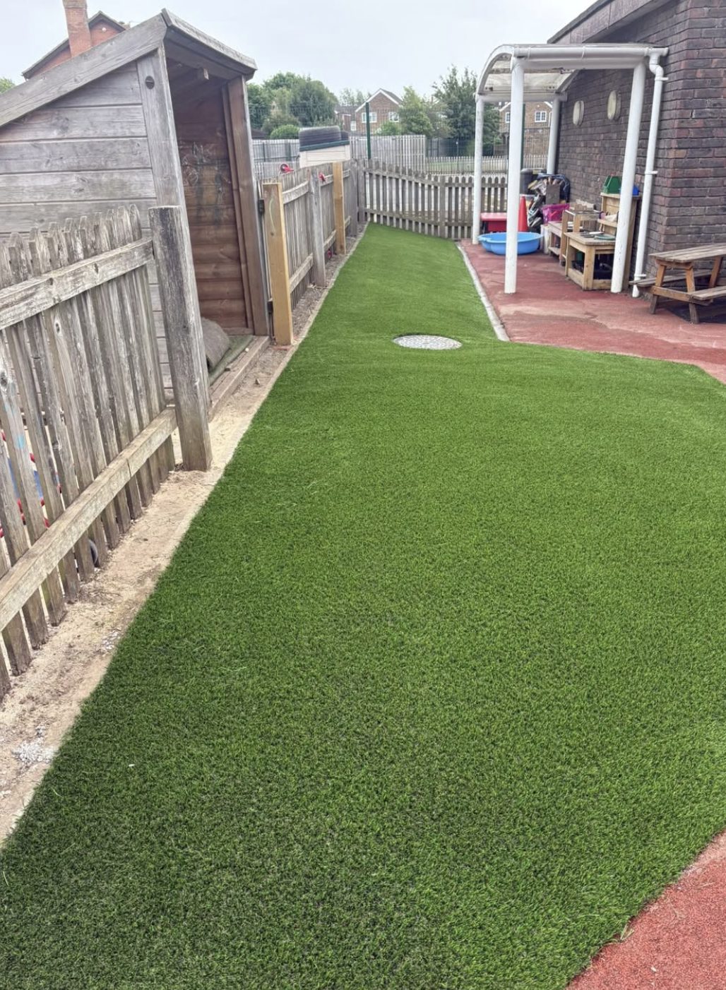 Artificial grass through a nursery play area between timber sheds and fencing, meeting red safety surfacing