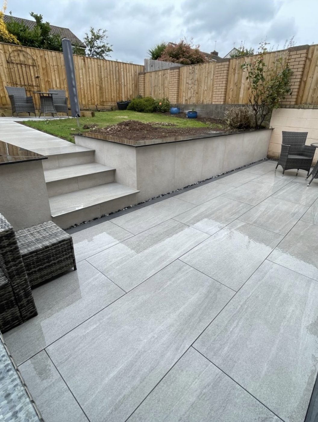 Grey porcelain patio with wide steps rising to a rendered retaining wall and upper lawn